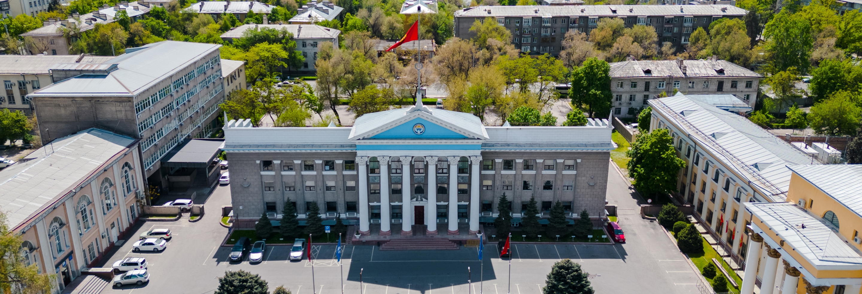 Здание с колоннами и голубой крышей в центре Бишкека, Киргизстан, окруженное зелеными деревьями и парковкой.